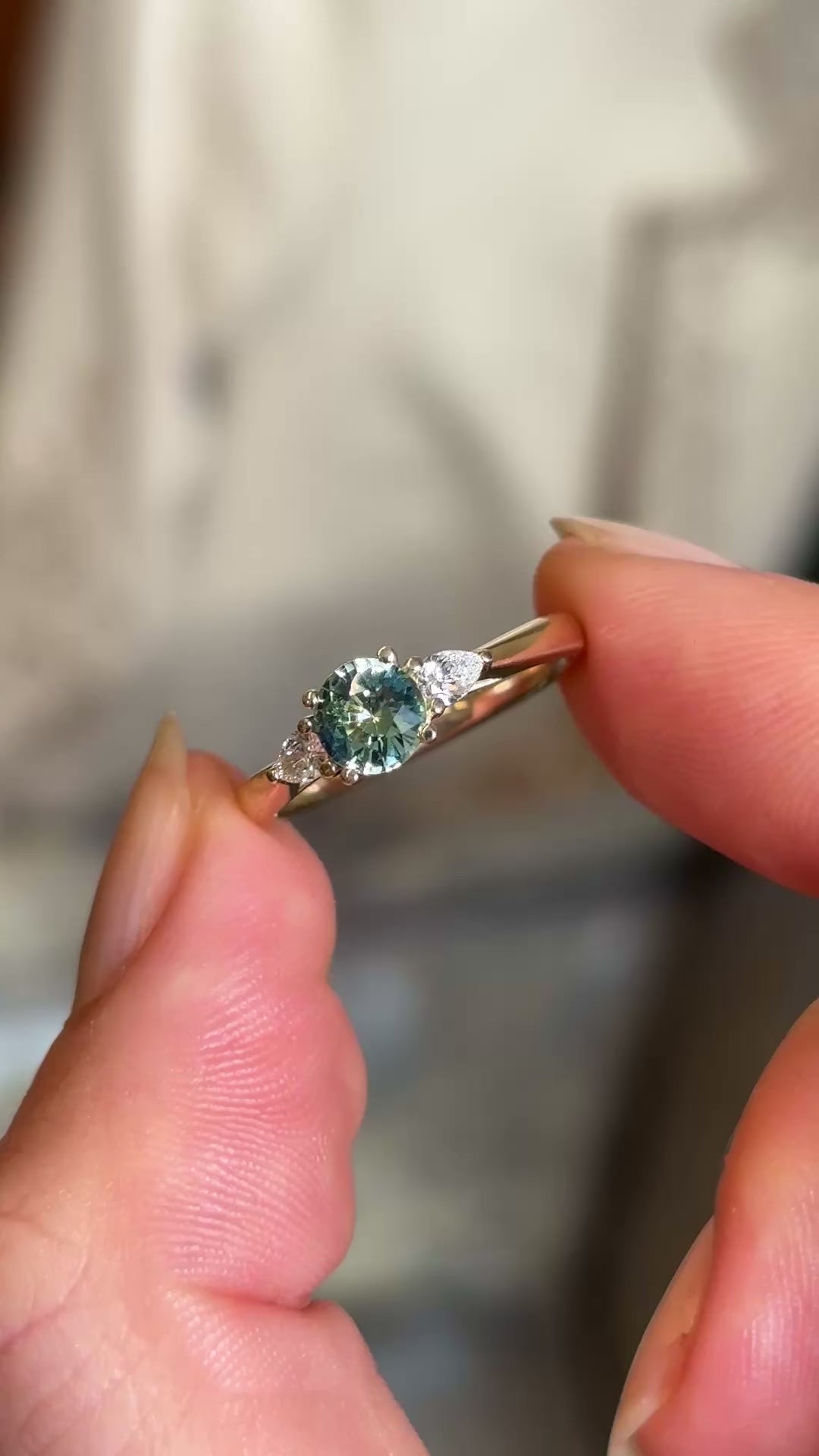 Round teal sapphire and pear-cut diamond three-stone engagement ring captured in natural light, emphasising the sparkle and brilliance of the gemstones.