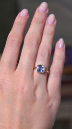 A video of an antique-inspired blue sapphire solitaire ring in a yellow gold bezel setting worn on the hand  against a blurry background.