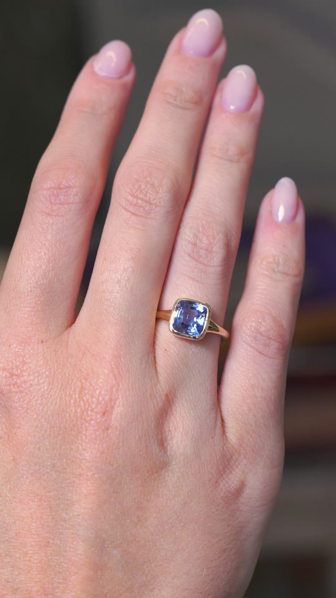 A video of an antique-inspired blue sapphire solitaire ring in a yellow gold bezel setting worn on the hand  against a blurry background.