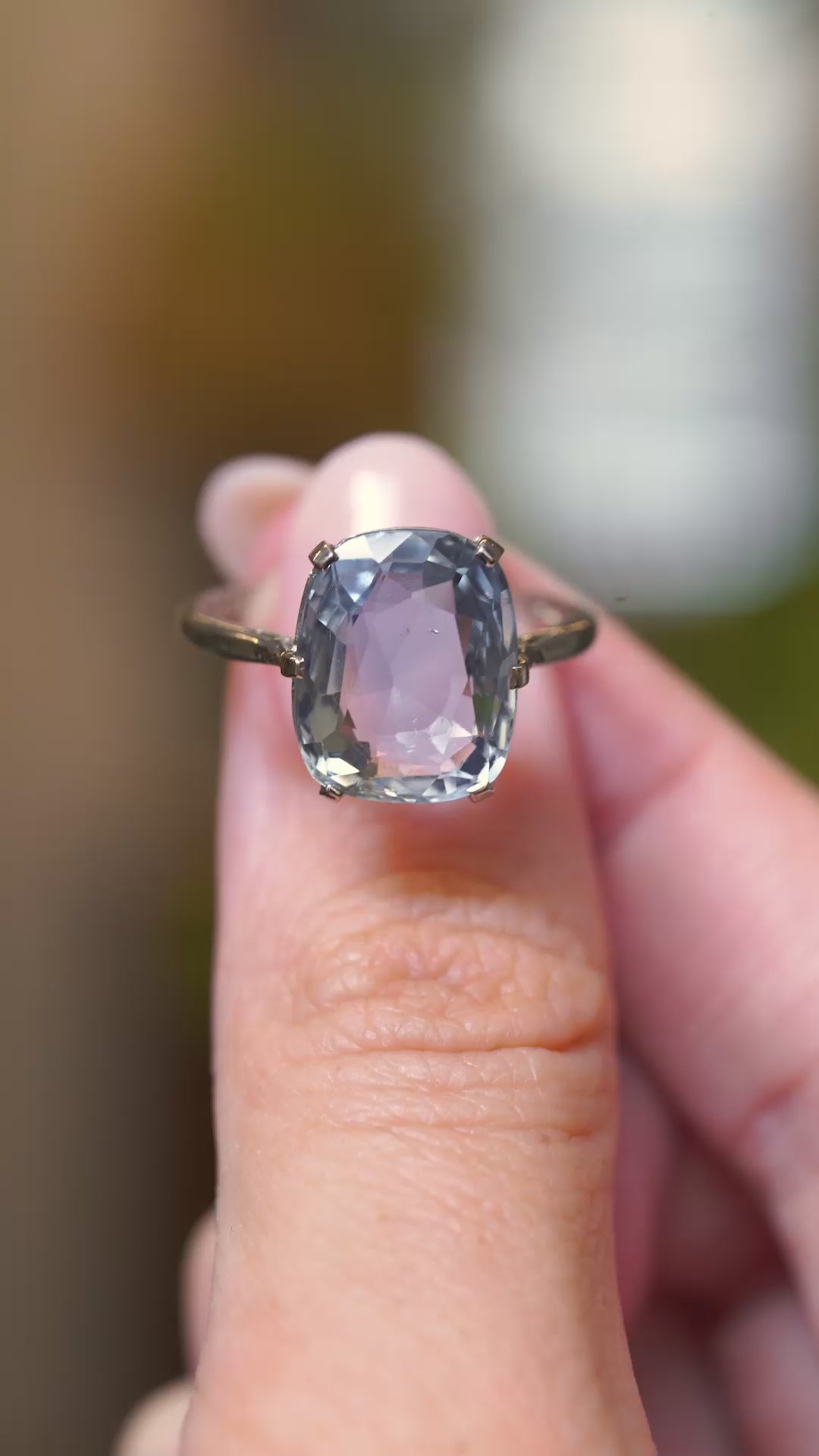 A video of a sapphire solitaire ring held in between fingers and turned to show various angles of the ring against a blurry background.
