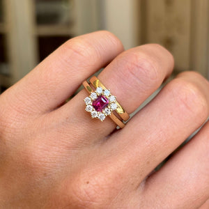 A cluster engagement ring featuring a rectangular step-cut ruby centre stone and sparkling diamond halo, worn on a finger with a gold wedding band.