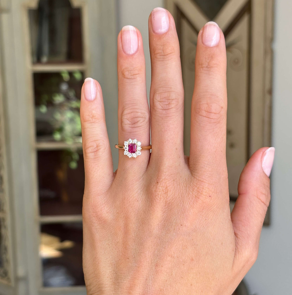 Rectangular step-cut ruby and diamond cluster engagement ring with a rectangular step-cut ruby centre stone and diamond halo, displayed on a hand.