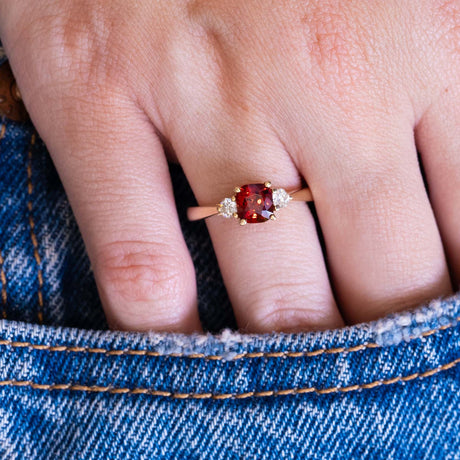Red spinel and diamond three-stone engagement ring worn on hand with denim pocket styling.