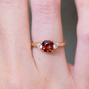 Red spinel and diamond three-stone engagement ring with cushion-cut centre stone worn on finger.