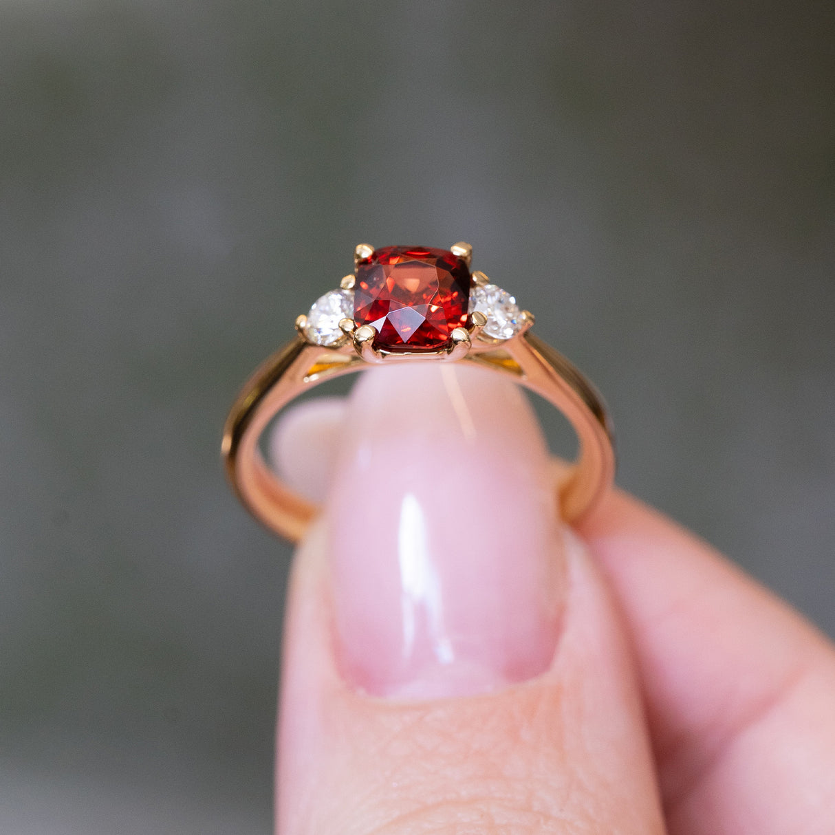 Red spinel and diamond three-stone engagement ring held between fingers with gold band.