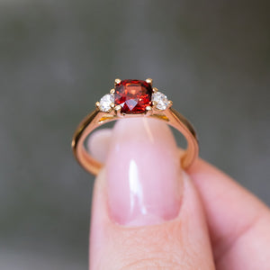 Red spinel and diamond three-stone engagement ring held between fingers with gold band.