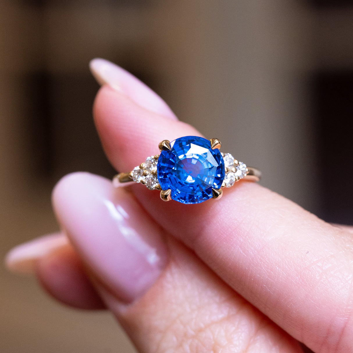 Round blue sapphire and diamond cluster tre-foil engagement ring held between fingers with brilliant blue sapphire centre stone.