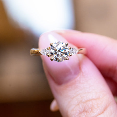 Round brilliant-cut diamond three-stone engagement ring held between fingers.