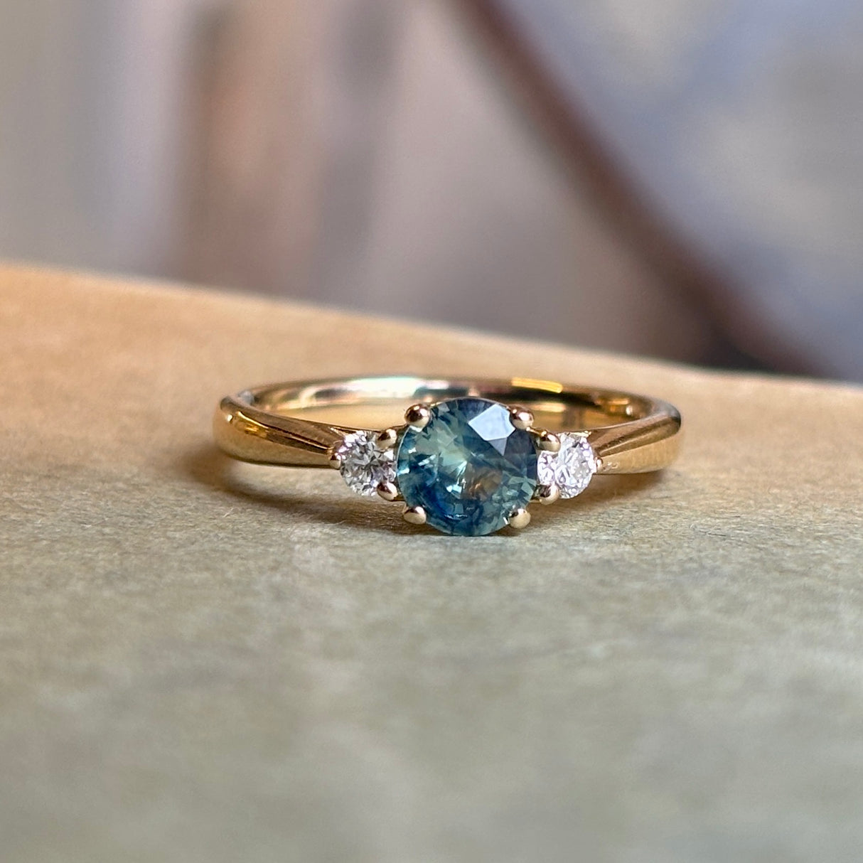 Round teal-green sapphire engagement ring with diamond side stones in polished yellow gold.