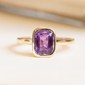 Royal purple cushion-cut sapphire solitaire engagement ring in a yellow gold rub-over setting.