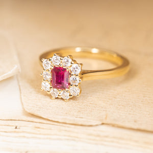 Rectangular step-cut ruby engagement ring with a diamond cluster surround and elegant yellow gold band.