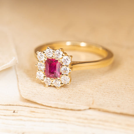 Rectangular step-cut ruby engagement ring with a diamond cluster surround and elegant yellow gold band.