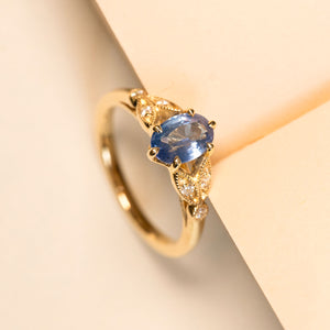 Yellow gold engagement ring featuring an oval blue sapphire framed by intricate diamond-set yellow gold shoulders.