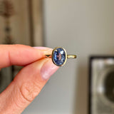 Antique-inspired oval-cut slate blue sapphire in a smooth yellow gold rub-over setting, held between fingers against a softly blurred vintage interior background.