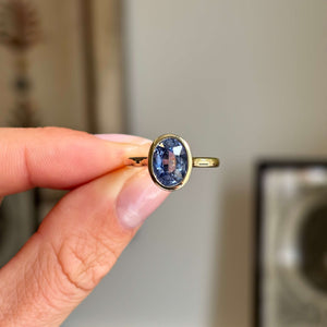 Antique-inspired oval-cut slate blue sapphire in a smooth yellow gold rub-over setting, held between fingers against a softly blurred vintage interior background.