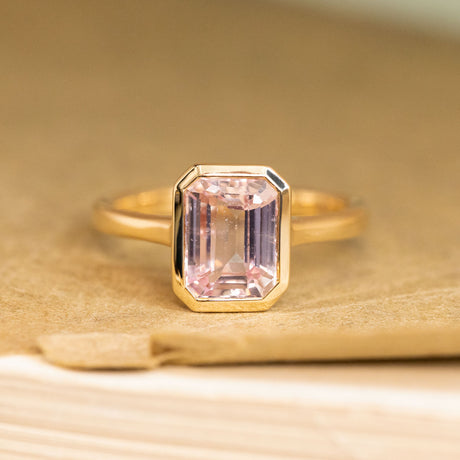 Soft petal-pink sapphire solitaire engagement ring featuring an emerald-cut pink sapphire set in polished yellow gold.