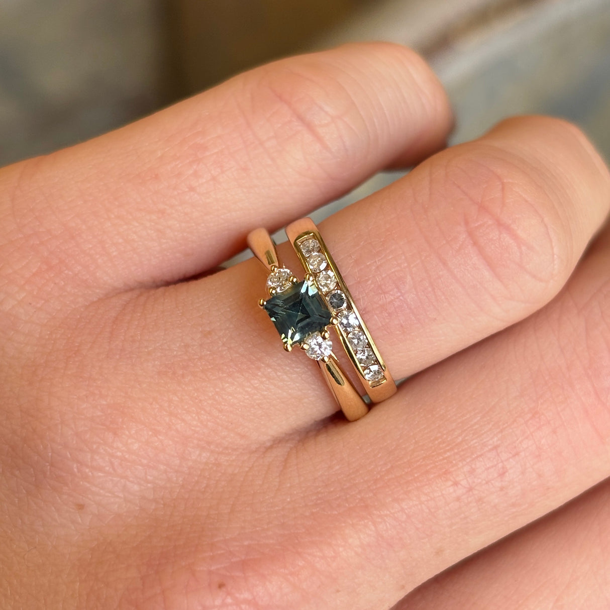 A square-cut teal sapphire (just under 1ct) and round-cut diamond three-stone engagement ring worn on the hand, showing its sparkle and design.