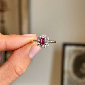 Antique-inspired step-cut pinkish-red ruby surrounded by brilliant diamonds in a yellow gold cluster setting, held between fingers against a softly blurred vintage interior background.