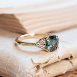 Teal sapphire and diamond cluster tre-foil engagement ring with diamond-set shoulders and yellow gold band.