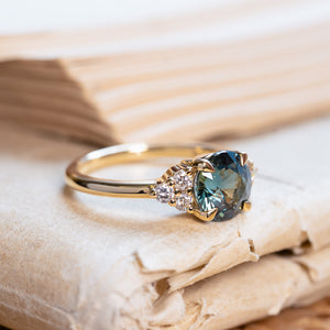 Teal sapphire and diamond cluster tre-foil engagement ring with diamond-set shoulders and yellow gold band.