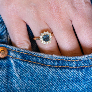 Teal sapphire and diamond cluster engagement ring worn with hand resting in denim pocket.