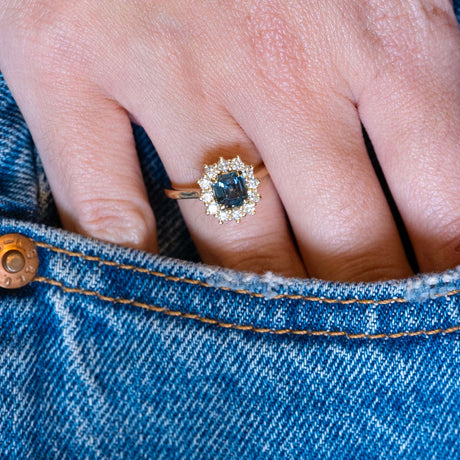 Teal sapphire and diamond cluster engagement ring worn with hand resting in denim pocket.