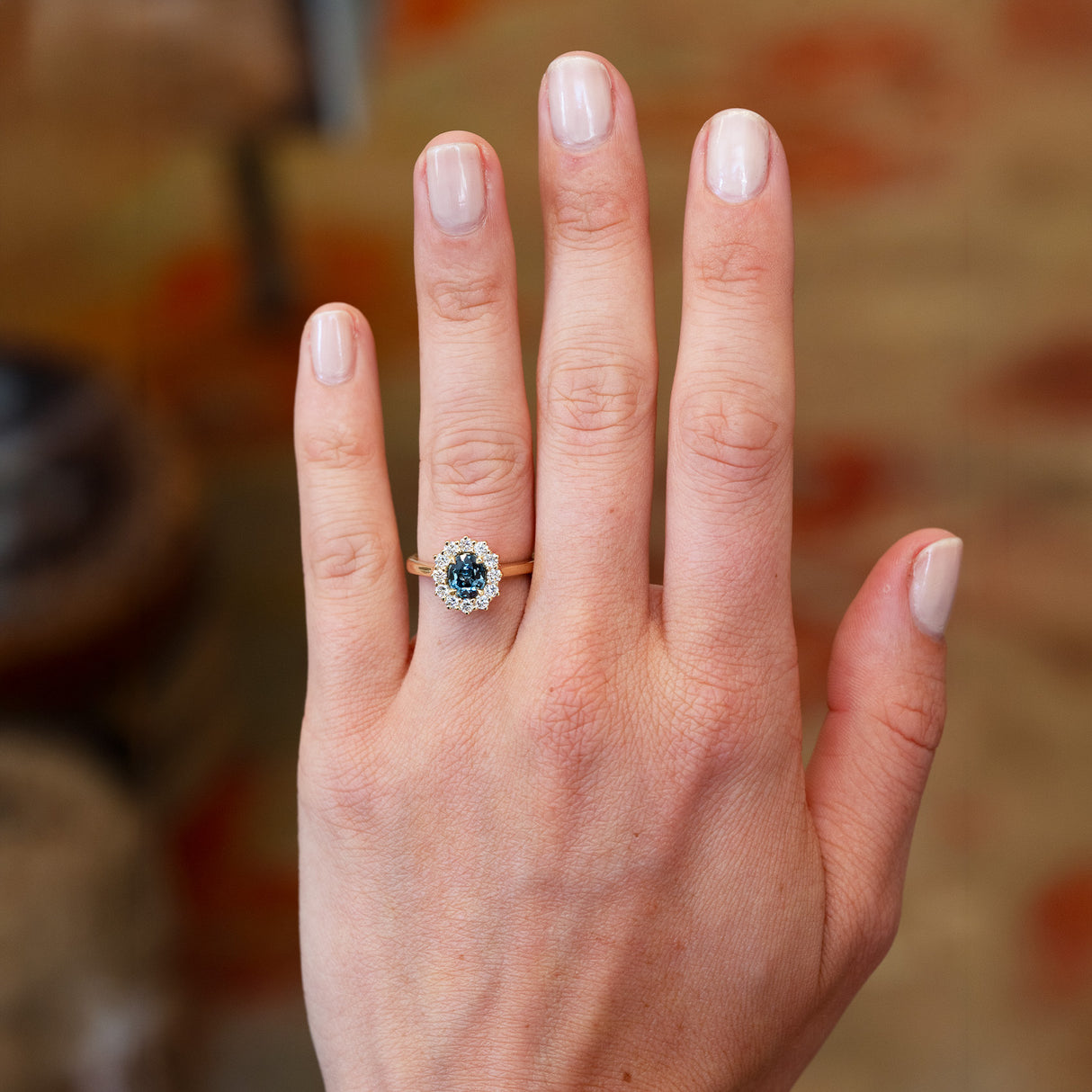 Teal sapphire and diamond cluster ring worn on hand with blurred background.