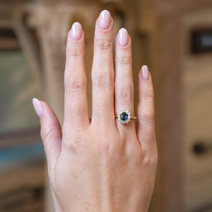 Teal sapphire and diamond cluster engagement ring worn on hand.