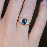 Close up of teal sapphire and diamond cluster trefoil engagement ring in yellow gold on a finger.