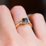 Teal sapphire and diamond three-stone engagement ring with gold band and diamond side stones.