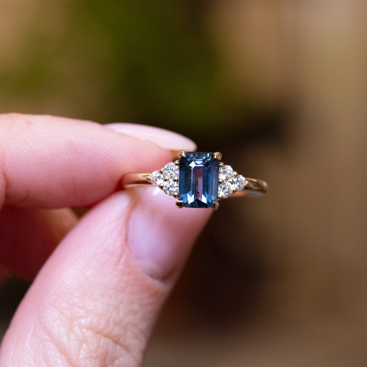 Teal sapphire and diamond cluster tre-foil engagement ring with geometric teal sapphire centre stone held between fingers.