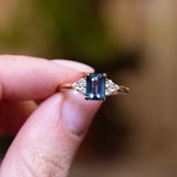 Teal sapphire and diamond cluster tre-foil engagement ring with geometric teal sapphire centre stone held between fingers.