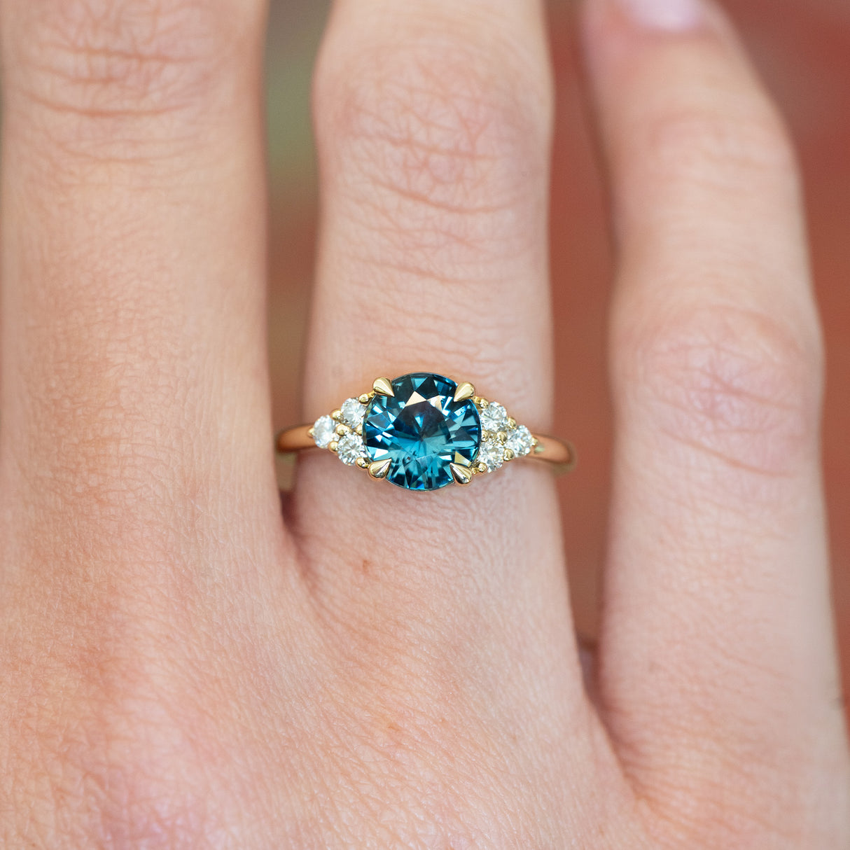 Teal sapphire and diamond cluster tre-foil engagement ring with diamond-set shoulders worn on finger.