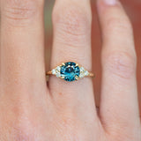 Teal sapphire and diamond cluster tre-foil engagement ring with diamond-set shoulders worn on finger.