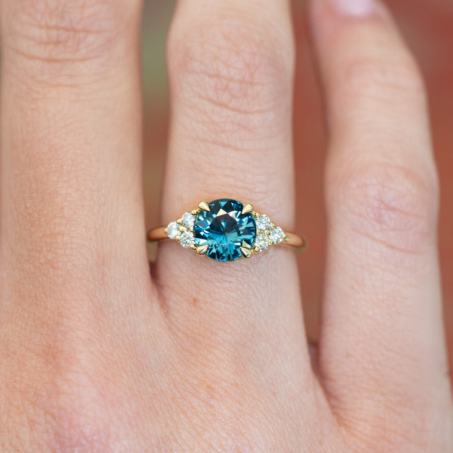 Teal sapphire and diamond cluster tre-foil engagement ring with diamond-set shoulders worn on finger.