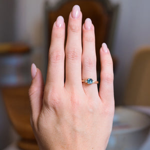 Teal sapphire and diamond three-stone engagement ring worn on hand with gold band.