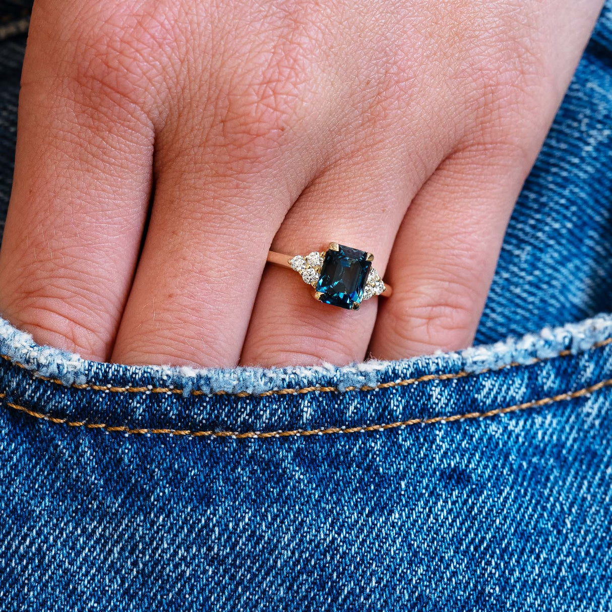 Close-up of a hand wearing a ring with a teal sapphire against a denim background.