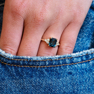Close-up of a hand wearing a ring with a teal sapphire against a denim background.