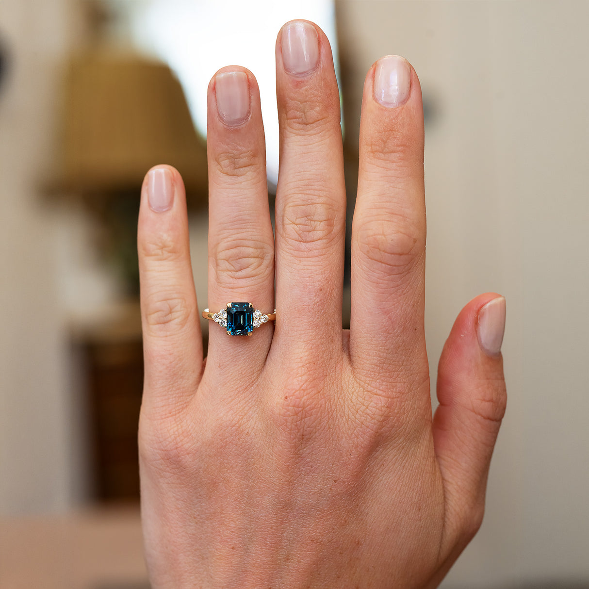 Teal sapphire and diamond ring worn on hand with blurred background.