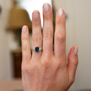 Teal sapphire and diamond ring worn on hand with blurred background.