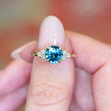 Teal sapphire and diamond cluster tre-foil engagement ring held between fingers with round teal sapphire centre stone.