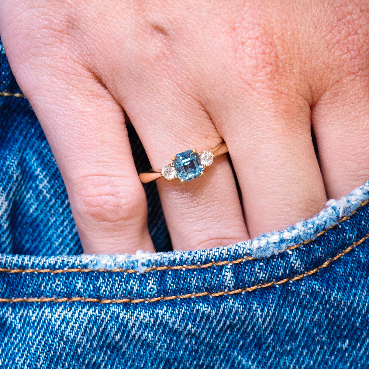 Teal sapphire and diamond three-stone engagement ring worn on hand with denim pocket styling.