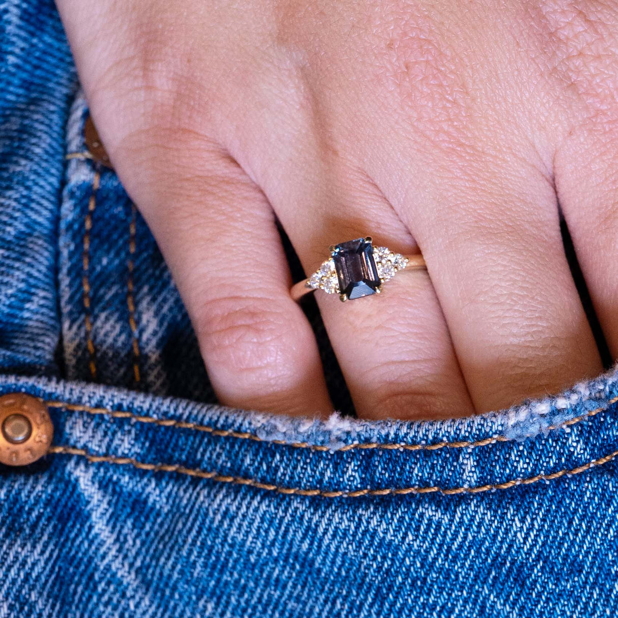 Teal sapphire and diamond cluster tre-foil engagement ring worn on hand with denim pocket styling.