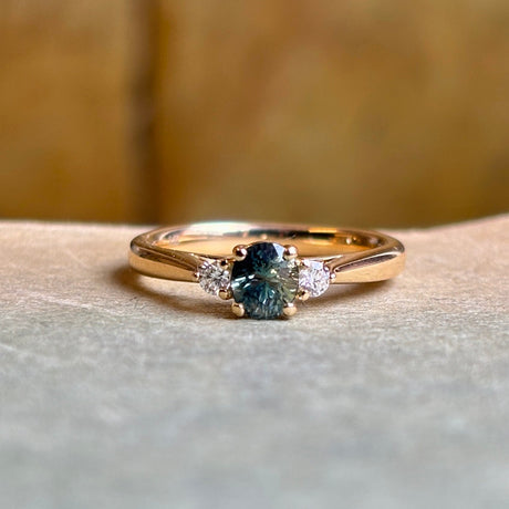 Angled view of teal sapphire engagement ring with diamond side stones in polished yellow gold.