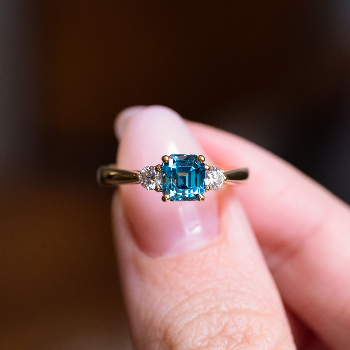Teal sapphire and diamond three-stone engagement ring held between fingers with emerald-cut teal sapphire centre stone.