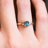 Teal sapphire and diamond three-stone engagement ring with rectangular teal sapphire centre stone worn on finger.