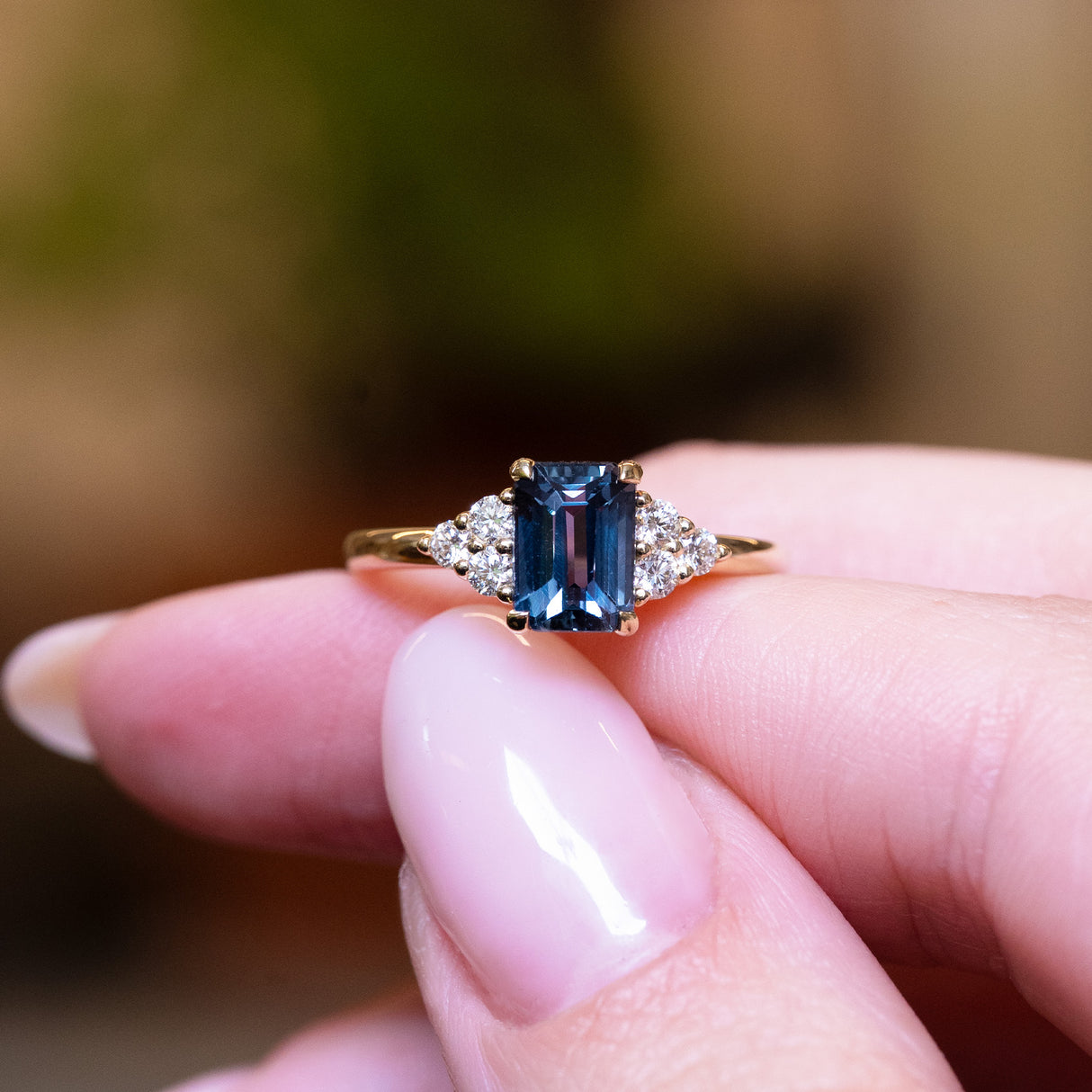 Teal sapphire and diamond cluster tre-foil engagement ring held on forefinger.