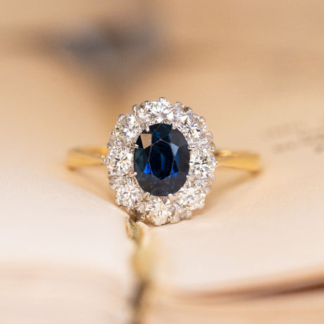 Vintage 1960s sapphire and diamond cluster ring featuring an oval-cut blue sapphire surrounded by a cluster of brilliant-cut diamonds in yellow gold.