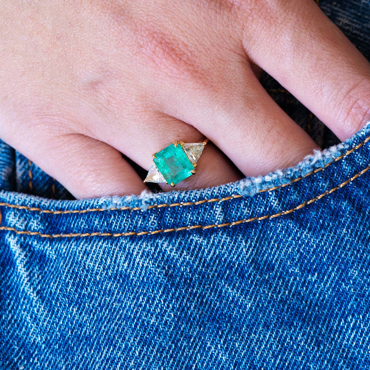 Close up of a vintage 1970s emerald and diamond three stone ring worn on hand with blue denim pocket detail.