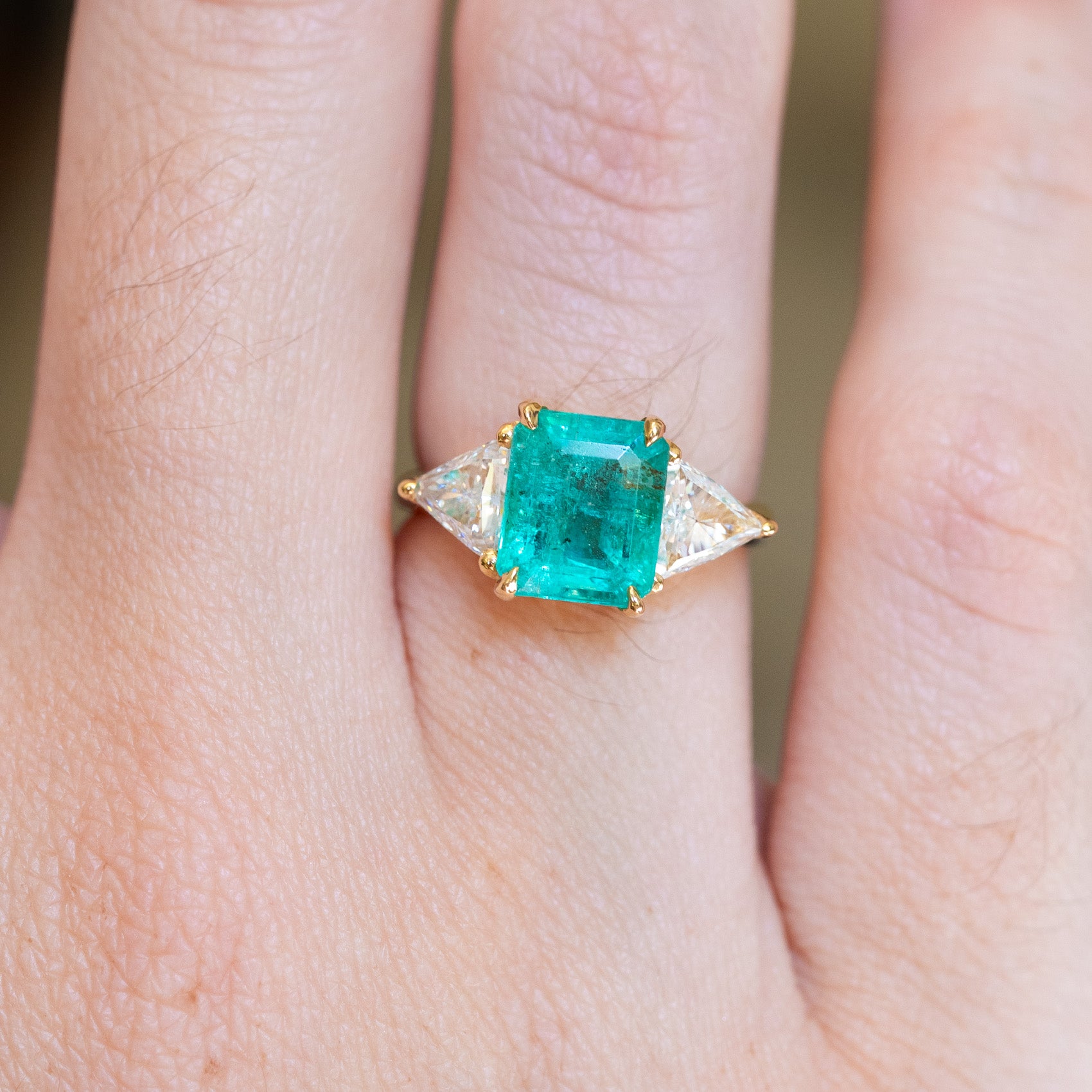 Emerald and diamond three stone ring with emerald cut centre and trilliant diamond side stones close up on hand.